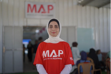 A Palestinian woman wearing a white hijab and a red MAP t shirt smiles at the camera.